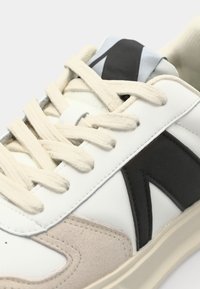 Białe skórzane sneakersy z kremowymi sznurowadłami, czarnymi i jasnoszarymi akcentami oraz miękką panelą z zamszu. Zauważalny detal przeszycia wzdłuż podeszwy.