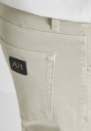 Lichtbeige denim broekzak met een zwart rechthoekig logoplaatje met gestileerde metalen letters "AM", dicht bij de bovenrand genaaid.