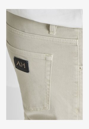 Tasca di pantaloni in denim beige chiaro con una toppa rettangolare nera con logo, cucita vicino al bordo superiore, che presenta le lettere stilizzate metalliche "AM".