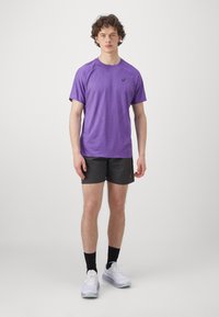 Männersport-T-Shirt in Lila mit kurzen Ärmeln, glatter Textur und dezentem Muster. Kombiniert mit schwarzen Shorts und weißen Turnschuhen.