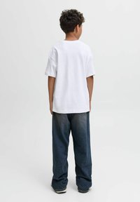 Un enfant aux cheveux bouclés porte un t-shirt blanc simple surdimensionné et un jean bleu foncé ample, se tenant de dos sur un fond blanc.