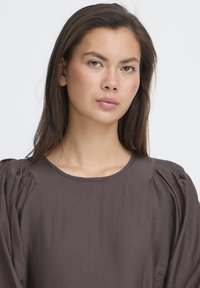 Brun tekstureret bluse med rund halsudskæring og pufærmer, der har et glat stof og diskret drapering. Ingen synlige mønstre eller hardware.