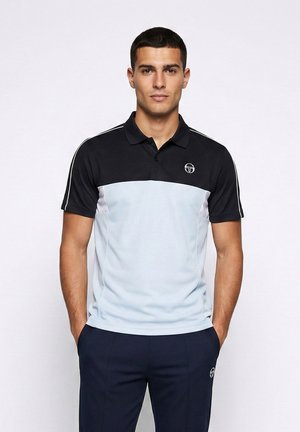 Jonge man staand met handen in de zakken, gekleed in een zwart en lichtblauw poloshirt met korte mouwen en donkerblauwe broek, tegen een witte achtergrond.