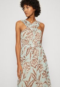 Il vestito con stampa floreale presenta uno sfondo azzurro chiaro con motivi botanici marroni. Le spalline incrociate mettono in risalto il décolleté e la vita.