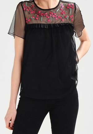 Czarna przezroczysta bluzka z krótkim rękawem z czerwonym kwiatowym haftem na piersi i falbaną poniżej dekoltu.