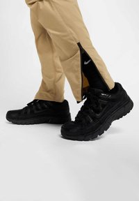 Czarne sportowe buty z teksturowaną siateczką i syntetycznymi nałożeniami, połączone z jasnobeżowymi spodniami z zamkami błyskawicznymi po bokach.