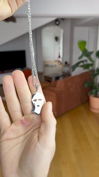 Ezüst maszk medál láncon, fényes felülettel, kivágott szemekkel és szájjal. A lánc apró, egymásba fonódó gyűrűkből áll.