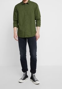 Grünes Langarmhemd mit kleinen weißen Punkten, Knopfleiste vorne und spitzen Kragen, kombiniert mit dunklen Jeans und schwarzen High-Top-Sneakern.
