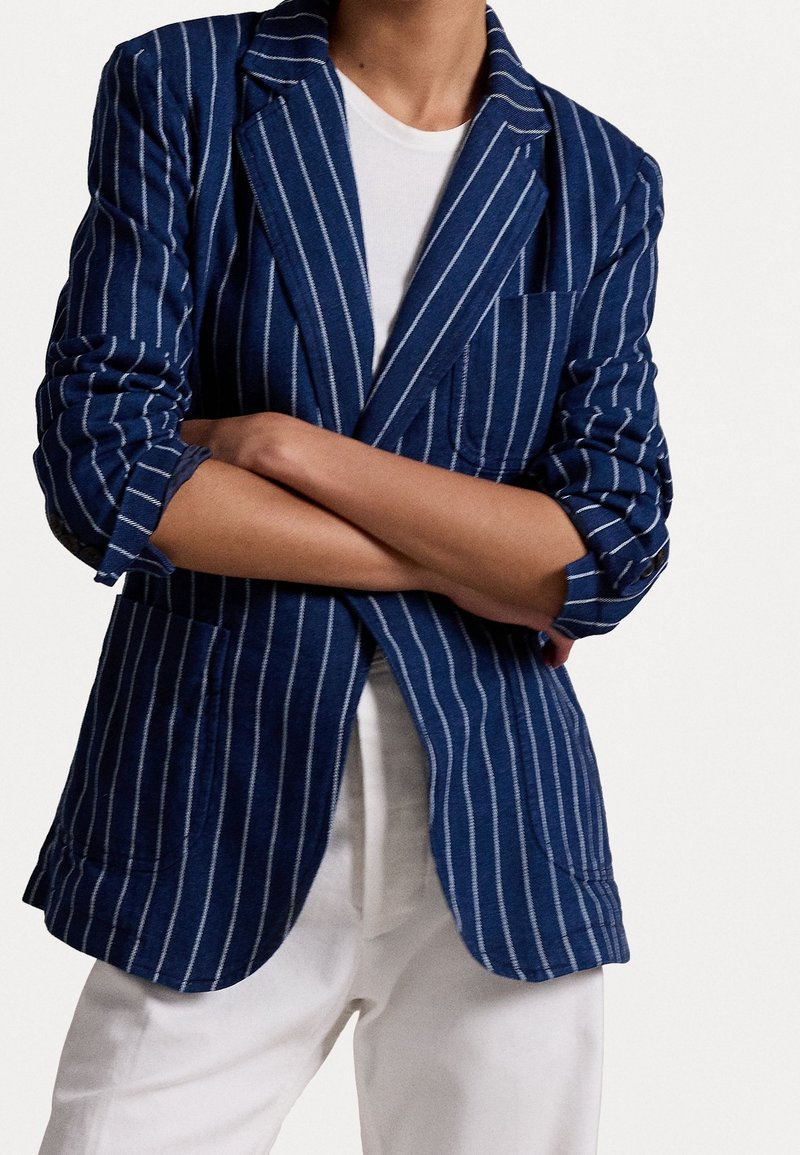 Blue and white striped blazer with notched lapels, front pockets, rolled sleeves, and a relaxed fit, paired with a white t-shirt.