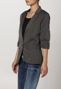 Grijze, op maat gemaakte blazer met een enkele knoopsluiting, gekartelde revers en opgerolde mouwen. Draagt over een zwart topje met blauwe jeans.