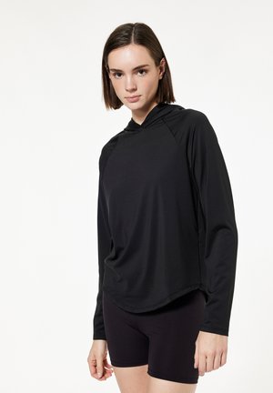 Jonge vrouw met kort bruin haar die een zwart hoodietop met lange mouwen en zwarte aangesloten shorts draagt tegen een witte achtergrond.