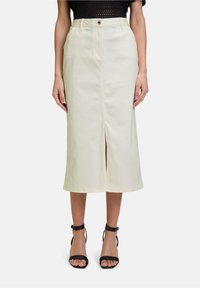 Jupe en denim beige longueur genoux avec une fente à l'avant, dotée d'une fermeture par bouton et de passants de ceinture, associée à des sandales à bride noire.