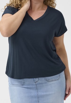 Osoba z kręconymi włosami, ubrana w granatowy T-shirt z dekoltem w serek i jasnoniebieską spódnicę dżinsową, z jedną ręką dotykającą szyi.