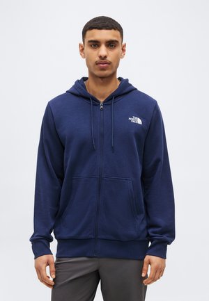 Hombre joven de pie mirando hacia adelante, vistiendo una sudadera con capucha azul marino con cremallera y el logo de The North Face en el pecho, y pantalones grises.