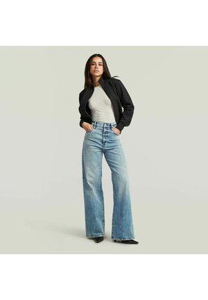 Vrouw staand met handen in zakken, gekleed in een zwart jack, lichtgrijs topje, wijde blauwe jeans en zwarte puntige schoenen tegen een effen achtergrond.