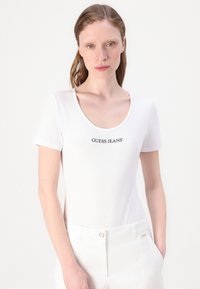 Camiseta blanca de manga corta con escote redondo y logo "GUESS JEANS" en negro a lo largo del pecho, fabricada con un tejido suave y liso.