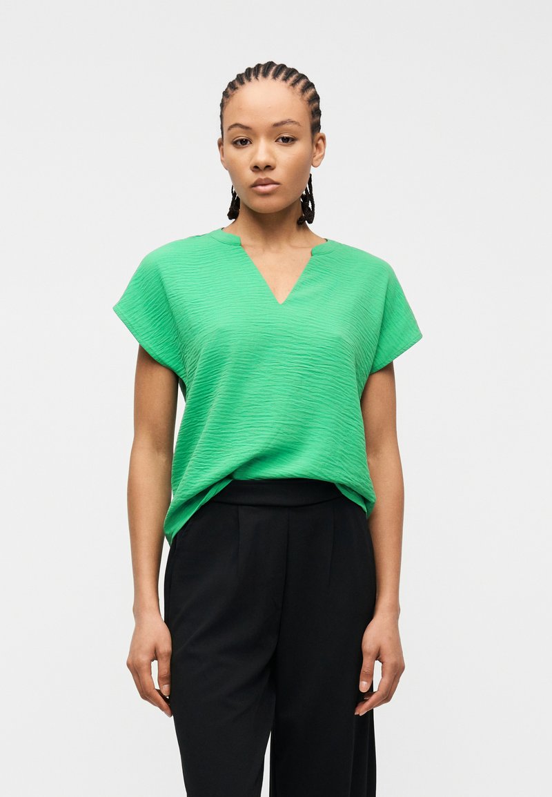 Γυναίκα με πλεξούδες που φοράει πράσινο μπλουζάκι με υφή και κοντά μανίκια, καθώς και ψηλόμεσο μαύρο παντελόνι, στέκεται μπροστά σε απλό φωτεινό φόντο.