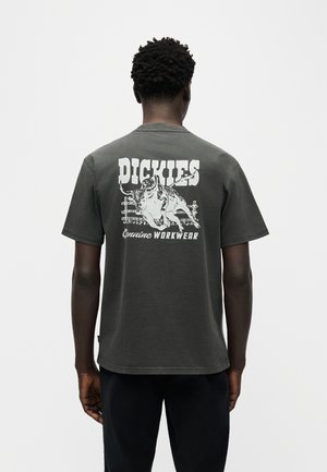 Ember szemből elfordulva, sötétszürke Dickies pólót visel, amelyen fehér bika grafika és a "Genuine Workwear" felirat található a hátoldalon.