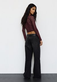 Hosszú sötét hajú nő, aki egy testhezálló bordó csipkés felsőt visel, laza fekete farmernadrággal, kissé oldalra fordulva áll egy fehér háttér előtt.