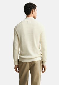 Strickpullover in Creme mit strukturiertem Muster, geripptem Kragen und Bündchen sowie einer taillierten Taille, kombiniert mit beigen Hosen.