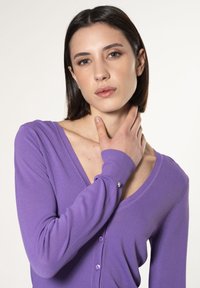 Cárdigan morado con escote en V, cierre de botones y mangas largas. Textura suave y diseño ajustado con un pequeño acento metálico.