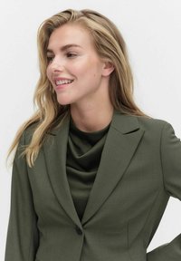 Olivgrüner Blazer mit elegantem Design, ausgestattet mit einem Reverskragen und einem einzigen Knopf. Darunter sorgt ein drapiertes dunkelgrünes Oberteil für Textur.