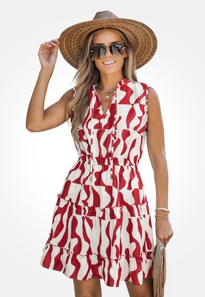 Femme portant une robe sans manches à motifs rouges et blancs, un chapeau de paille à large bord, de grandes lunettes de soleil et tenant une pochette beige.