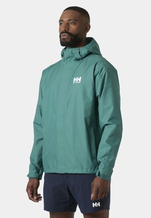 Man met een waterdicht teal Helly Hansen jack met capuchon en donkerblauwe korte broek tegen een effen achtergrond.
