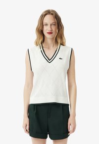 Gilet bianco senza maniche a maglia con scollatura a V, caratterizzato da un motivo a diamante e rifiniture verde scuro, abbinato a pantaloni corti verde scuro.