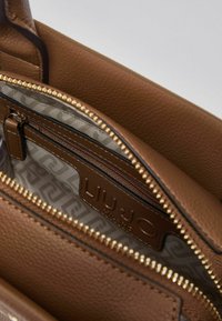 Intérieur d'un sac à main en cuir marron avec une poche zippée, une fermeture éclair dorée et une étiquette de marque. La doublure présente un motif rayé.