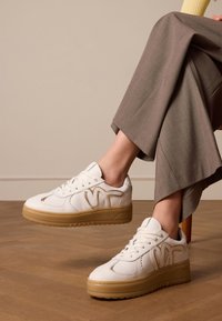 Witte leren sneakers met textuuraccenten, voorzien van een dikke gumrubberen zool en gestileerd zijlogo. Draagt een combinatie met bruin wijde broek.