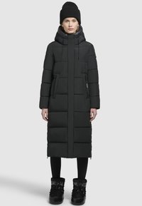 Schwarze Daunenjacke mit knielangem Schnitt, mit Reißverschluss vorne, Kapuze mit Kordelzug und Seitentaschen. Trägt dazu schwarze Leggings und Stiefel.