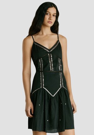 Jonge vrouw met lang donker haar, gekleed in een zwarte mouwloze jurk met zilveren paillettenversieringen op het lijfje en de rok, staand tegen een grijze achtergrond.