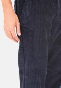 Pantaloni in corduroy blu navy con righe verticali, dotati di tasche laterali e una superficie liscia. Posizionati a mano vicino alla cucitura del capo.