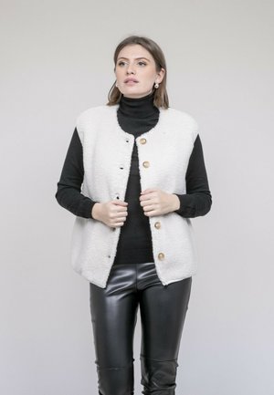 Femme aux cheveux châtain clair portant un gilet en polaire blanc boutonné sur un col roulé noir et un pantalon en cuir noir, debout devant un fond gris clair.