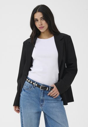 Vrouw met lang donker haar, gekleed in een zwarte blazer, witte top, blauwe spijkerbroek en zwarte gesp met studs, staand tegen een effen achtergrond.