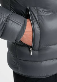 Dettaglio di un piumino grigio scuro che mostra una tasca con zip e un tirante argento, oltre a polsini a coste. Texture liscia e imbottita visibile.