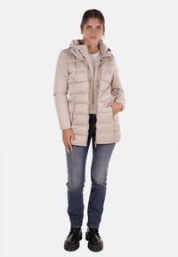 Giacca a vento leggera beige chiaro con cappuccio, chiusura a zip, texture trapuntata e tasche laterali, abbinata a jeans blu e stivali neri.