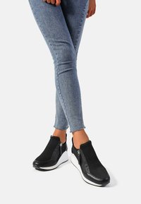 Sneakers neri slip-on con tomaia in tessuto testurizzato, suola a zeppa bianca e dettagli con cerniera sui lati, abbinati a jeans blu aderenti.