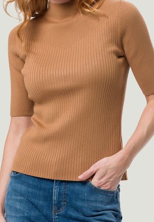 Kvinde iført en tætsiddende, ribbet, kamelbrun kortærmet sweater, der er let stoppet ned i blå cowboybukser, med den ene hånd i lommen.
