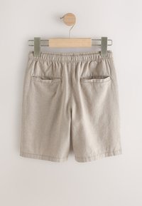 Pantalones cortos beige con cintura elástica y dos bolsillos traseros colgados en una percha de madera contra un fondo claro.