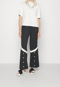 Kvinna som bär svarta vida byxor med vita snedstreck och knappar, tillsammans med en vit t-shirt och svart-vita sandaler.