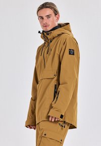Bruine, waterdichte jas met een capuchon, een voork pocket en ritszakken aan de zijkant. Voorzien van trekkoorden en branding op de mouw.