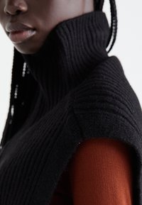 Nahaufnahme einer Person, die einen schwarzen gerippten Rollkragenpullover über einem rostfarbenen gerippten Oberteil trägt und geflochtenes Haar zeigt.