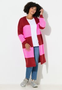 Cardigan lavorato a maglia con audaci righe orizzontali rosse e rosa, design con apertura frontale e maniche lunghe. Abbinato a jeans blu e sneakers argento.