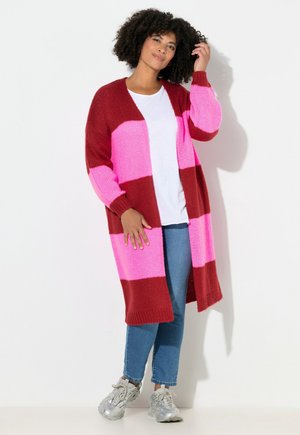 Strikcardigan med kraftige røde og pink horisontale striber, åben frontdesign og lange ærmer. Parret med blå jeans og sølvfarvede sneakers.
