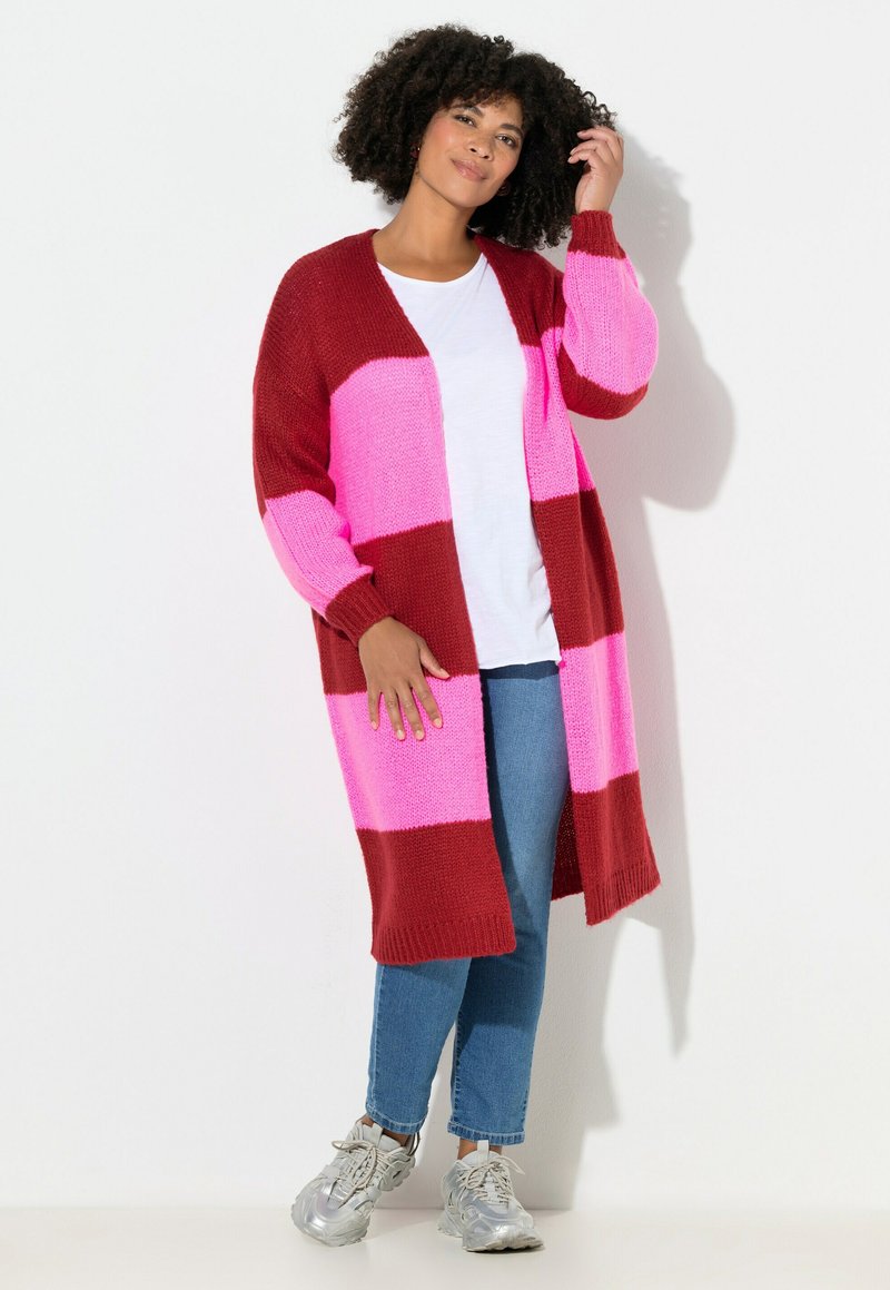 Cardigan lavorato a maglia con audaci righe orizzontali rosse e rosa, design con apertura frontale e maniche lunghe. Abbinato a jeans blu e sneakers argento.