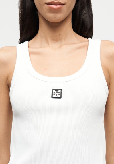 Λευκό ριπ τοπ με λογότυπο σε σχήμα γεωμετρικού σχεδίου στο κέντρο. Το ύφασμα έχει ομαλή υφή και εφαρμοστή γραμμή.