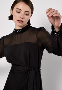 Vestido negro con una blusa larga y transparente. Cuenta con un cuello adornado con lentejuelas y una cintura ajustada, ofreciendo una silueta estructurada.