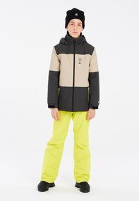 Skijacke mit beige und schwarzen Abschnitten, mit Reißverschluss und Logo; kombiniert mit leuchtend gelben Skihosen und schwarzen Stiefeln.